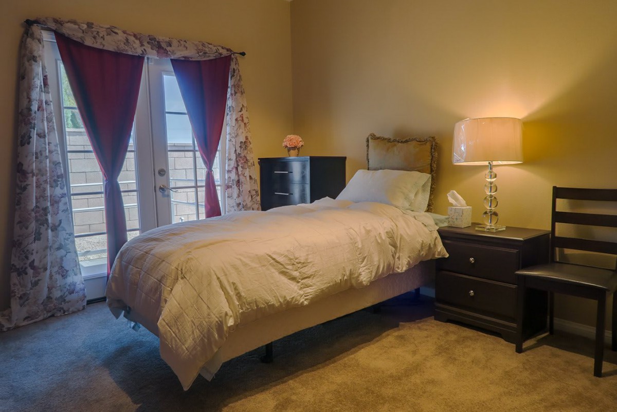A cozy bedroom with a single bed, nightstand, and lamp next to French doors covered by curtains.