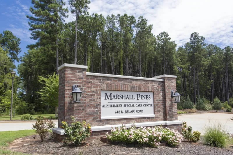 Entrance sign for Marshall Pines Alzheimer's Special Care Center, surrounded by trees and landscaping.