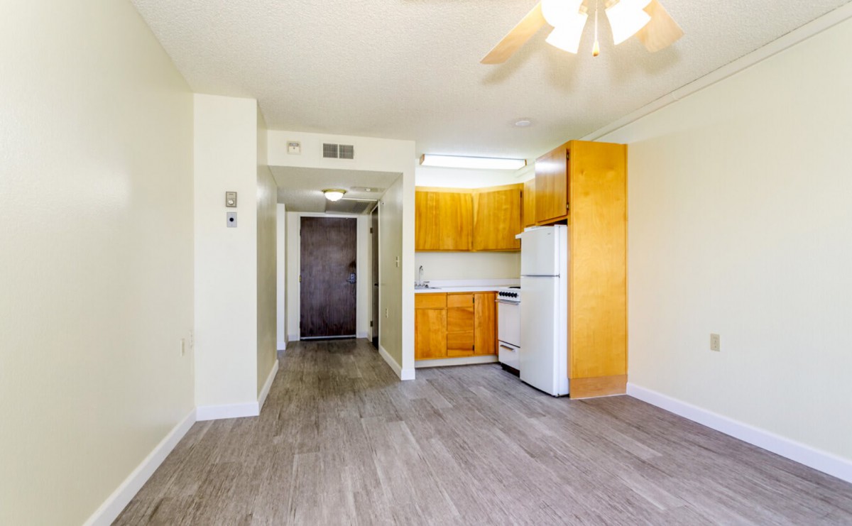A view of a small apartment kitchen with wooden cabinets and a refrigerator, featuring a ceiling fan and hardwood flooring.