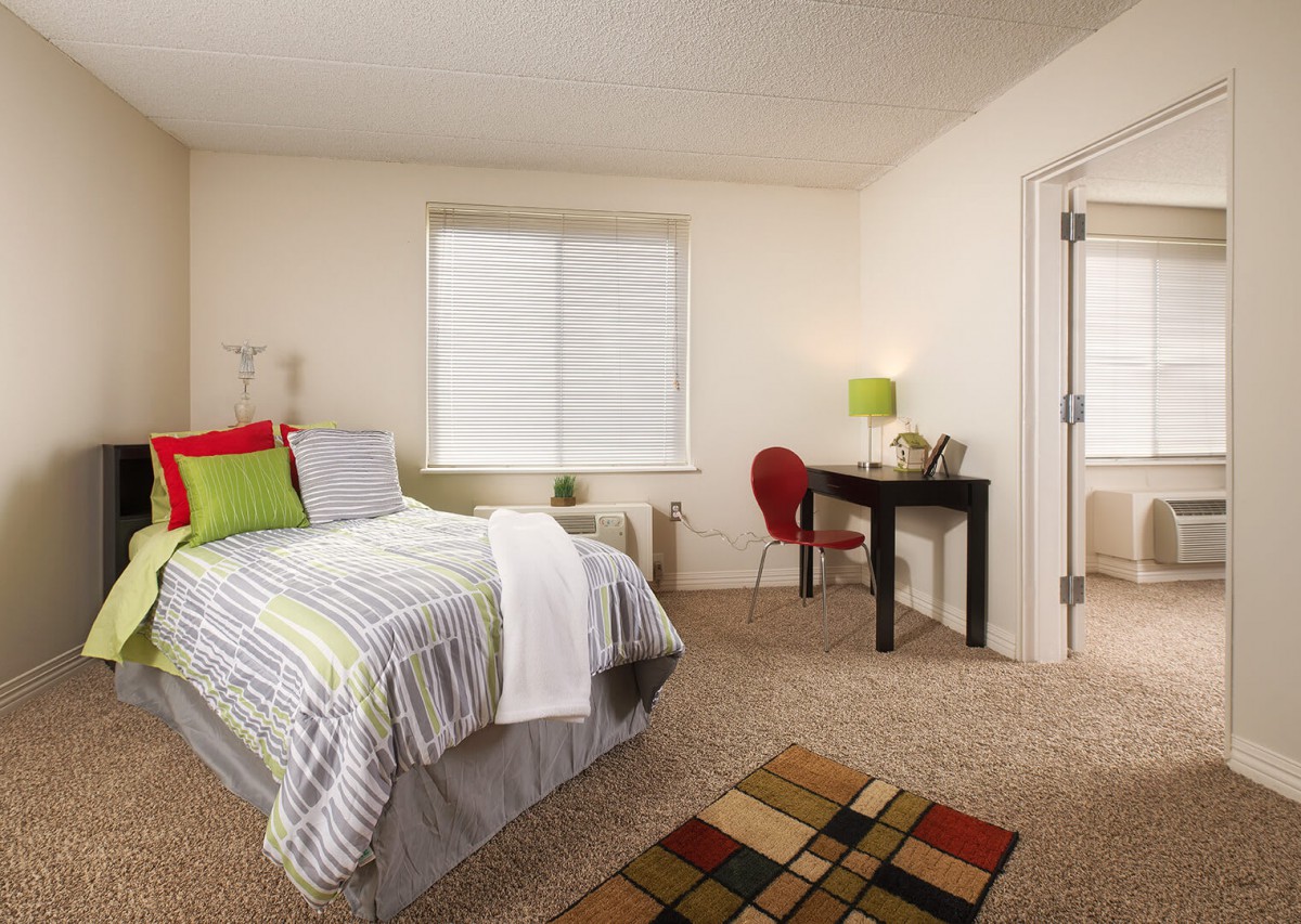 A cozy bedroom with a neatly made bed, a desk with a lamp, and a small carpet on the floor.