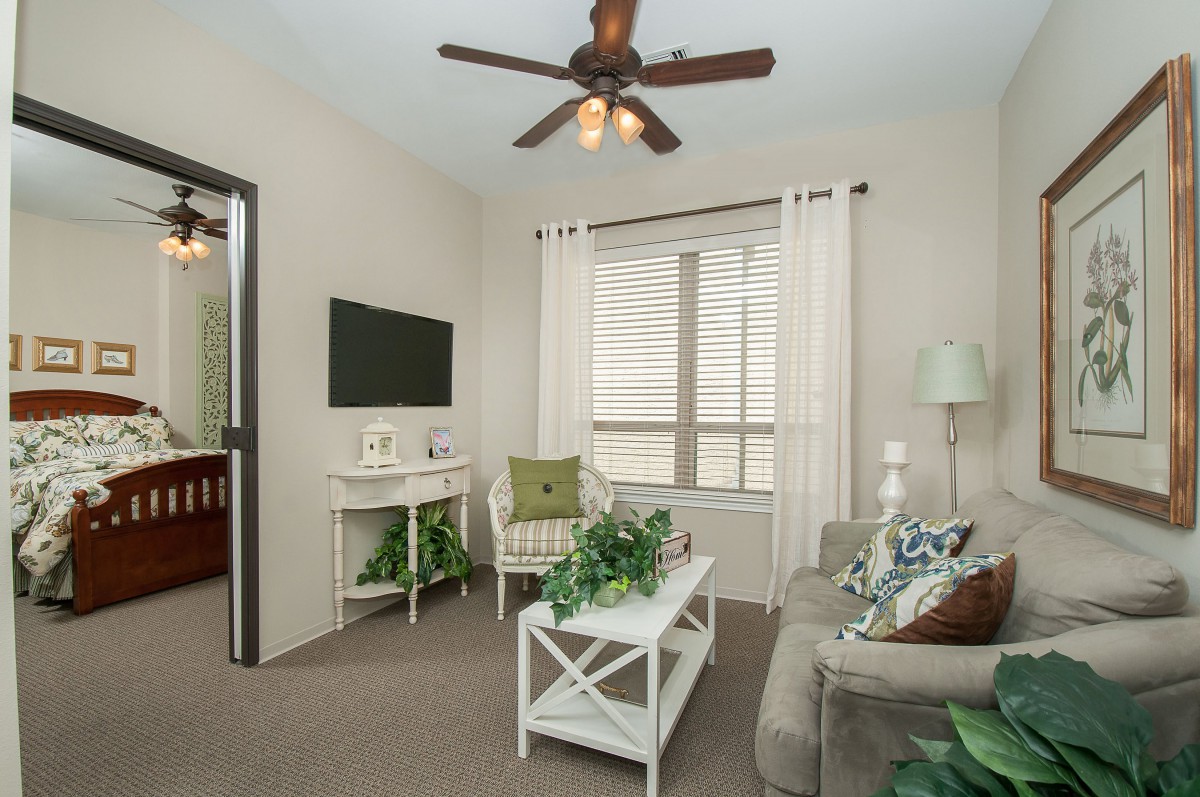 A cozy living room with beige walls, featuring a comfortable sofa, a white coffee table adorned with plants, and a wall-mounted TV, illuminated by natural light streaming through the window.