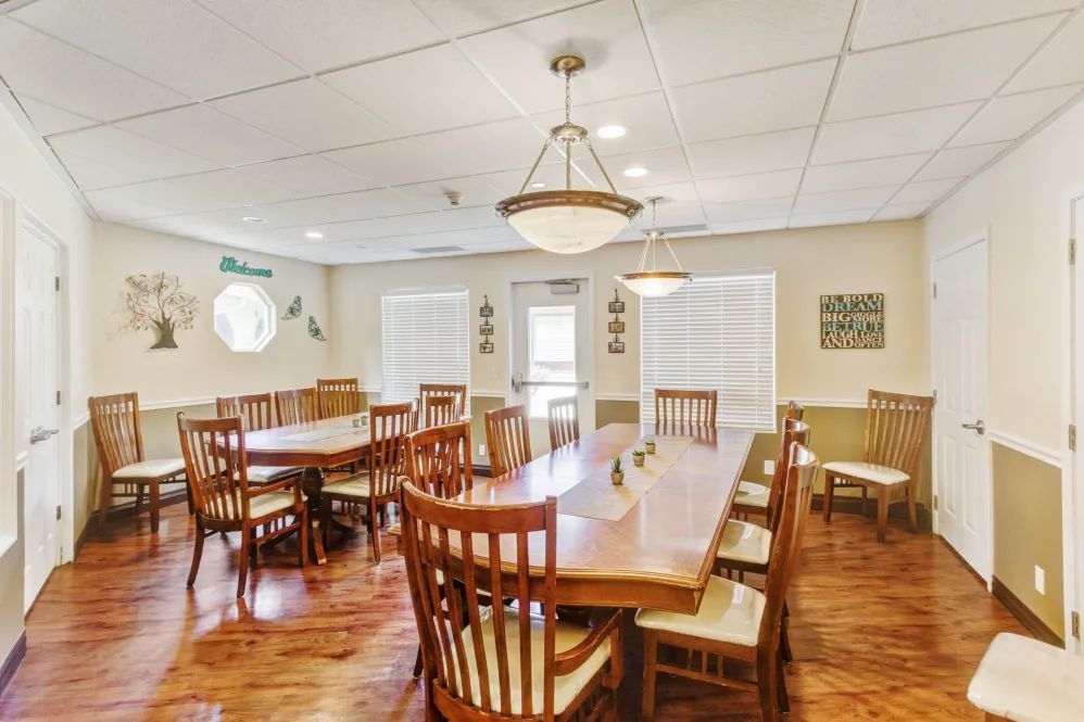 A spacious dining room with wooden tables and chairs, illuminated by overhead chandeliers, featuring natural light from large windows.
