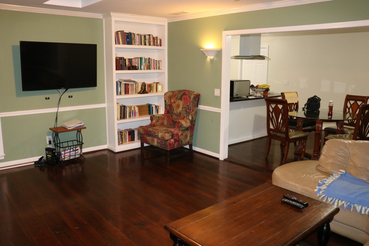 A cozy living room with hardwood floors, featuring a bookshelf, armchair, and wall-mounted TV, with an open view to the kitchen.