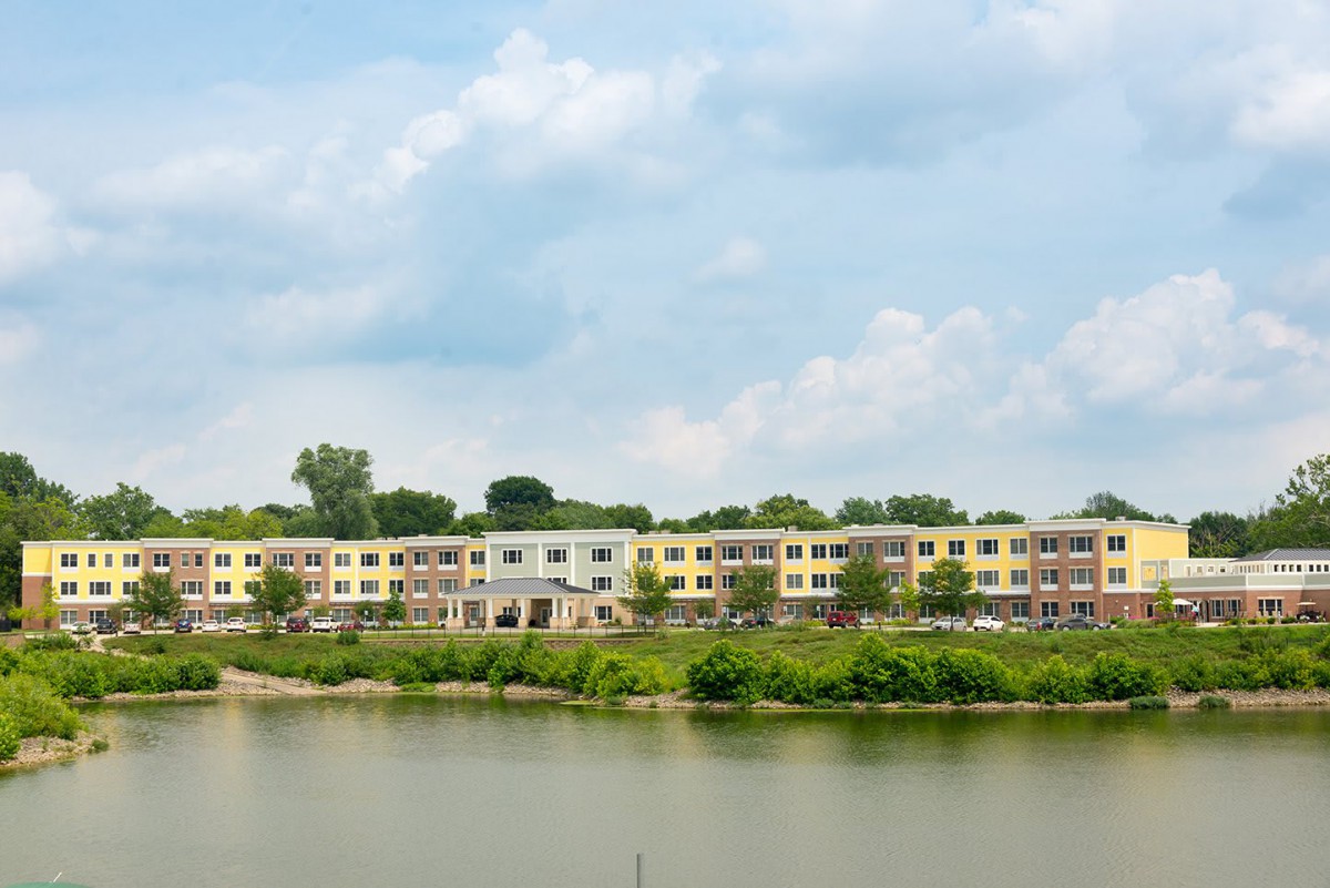 A row of residential buildings overlooking a calm body of water, surrounded by lush greenery under a partly cloudy sky.