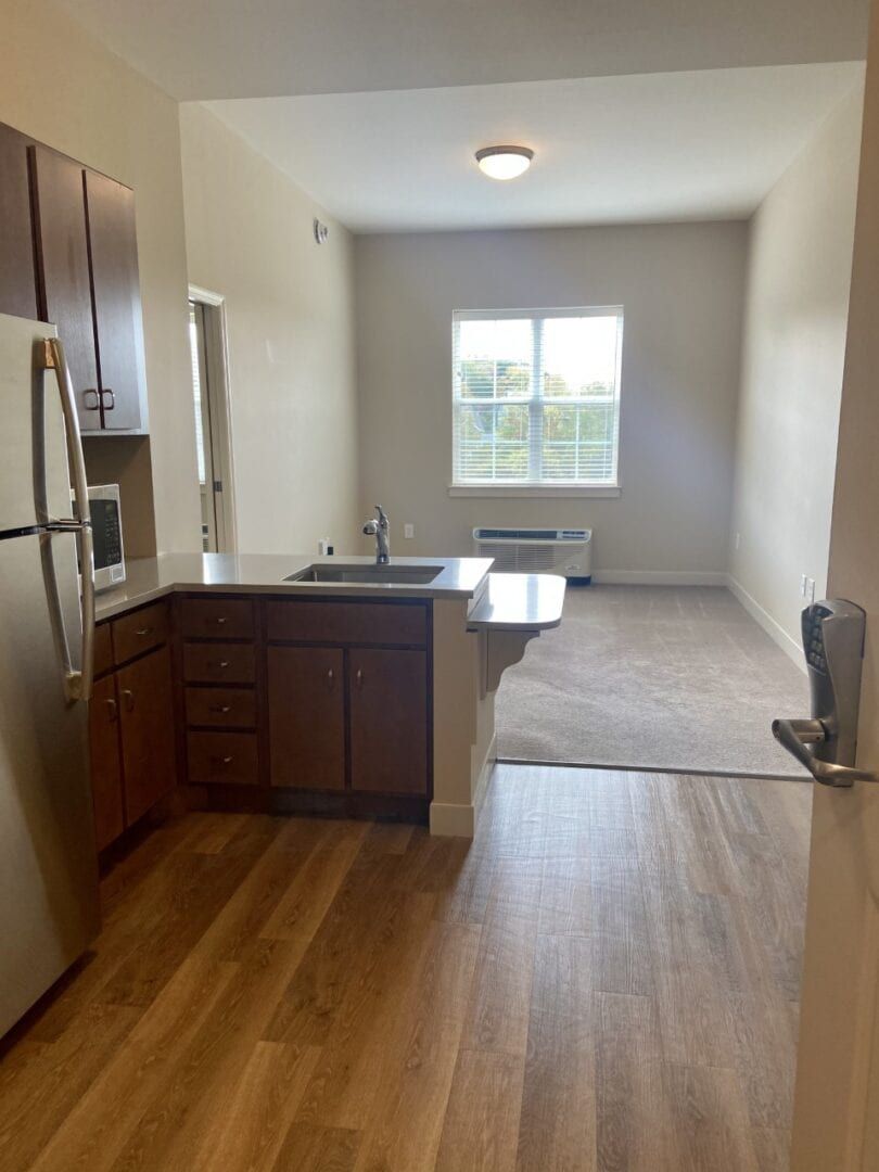 A view of an apartment with an open kitchen and living room area, featuring hardwood floors and a large window.