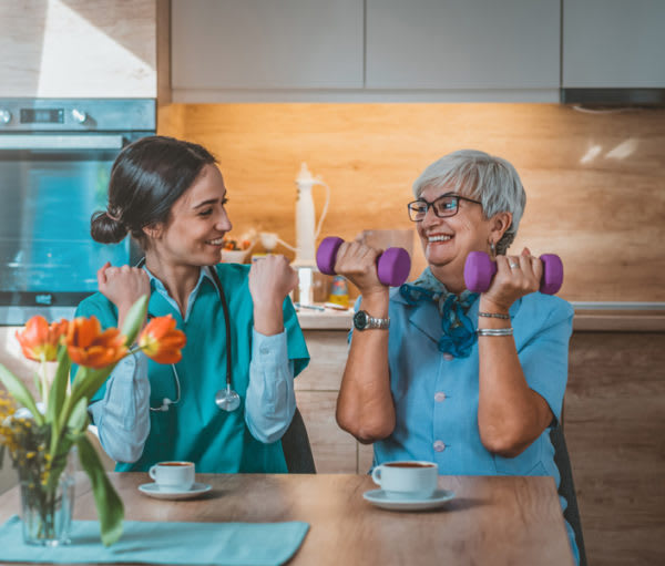 A healthcare worker and a senior woman exercising with dumbbells in a cozy indoor setting.