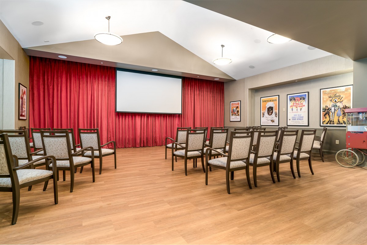 A small auditorium setup with chairs facing a projector screen and red curtains.