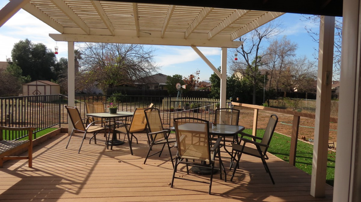 A sunny patio with tables and chairs under a pergola, overlooking a suburban yard.