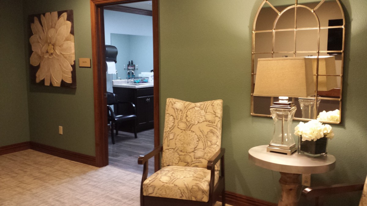 A cozy waiting area with a patterned chair, lamp, and decorative mirror near an open door leading to another room.