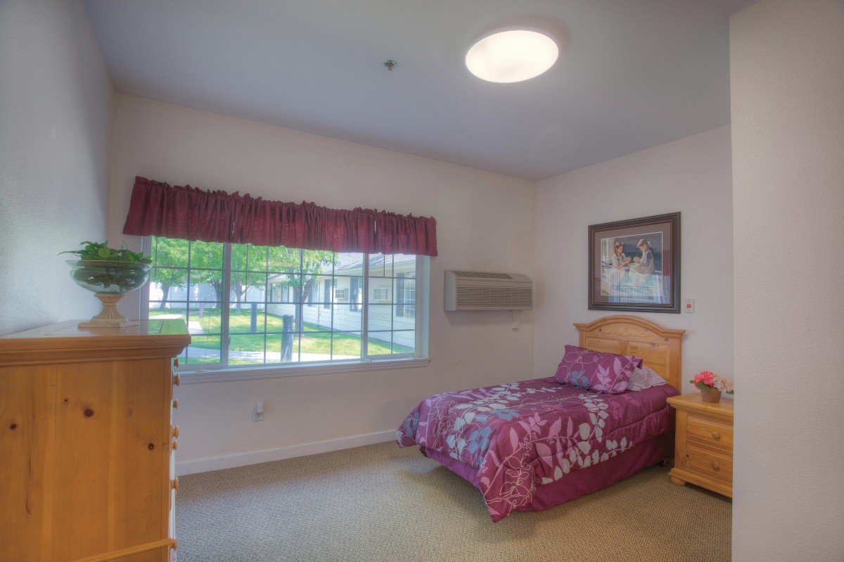 A cozy bedroom with a single bed, wooden furniture, and a large window overlooking greenery.