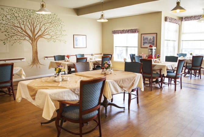 An empty dining room with tables and chairs arranged neatly, featuring wall art and bright natural light from the window.