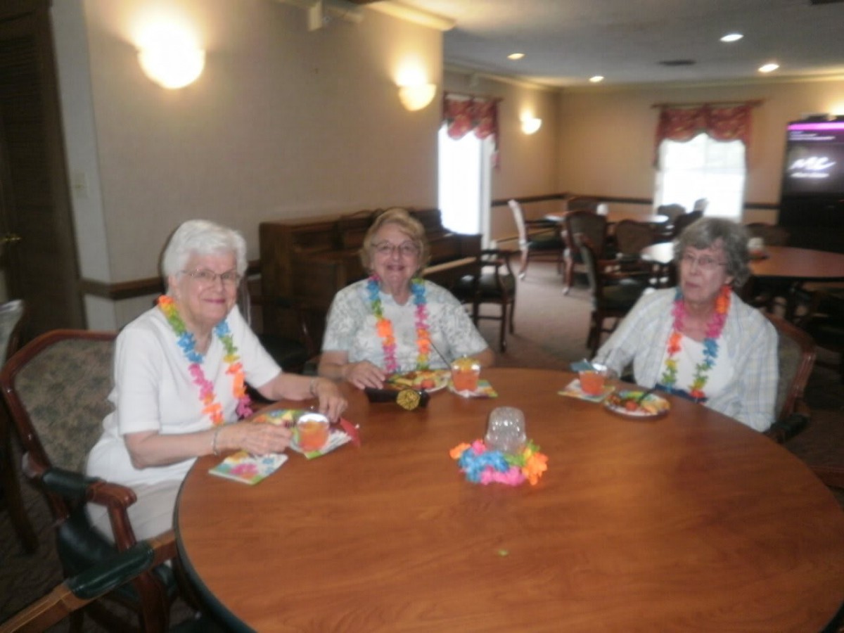 Three elderly women enjoying a meal together at a dining table, adorned with colorful leis.