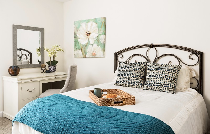 A cozy bedroom featuring a bed with patterned pillows, a teal blanket, and a decorative tray. The room is complemented by a floral painting and a mirror on the wall.