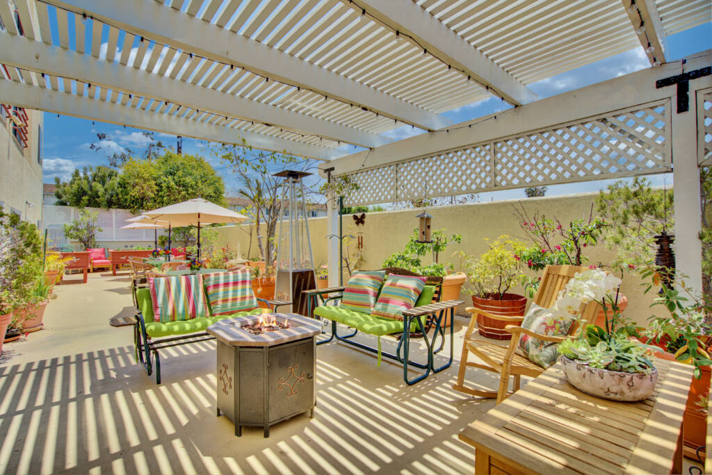 A sunny patio with a pergola, featuring comfortable outdoor seating and a fire pit surrounded by lush plants.