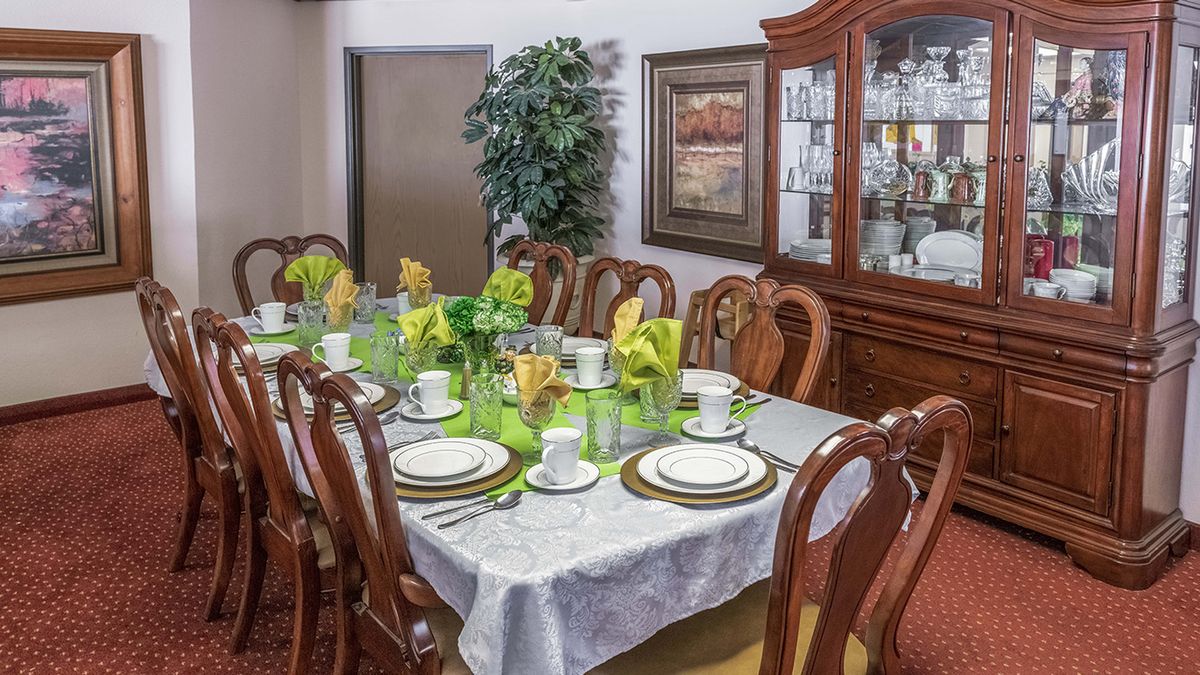 An elegantly set dining table with green and gold accents, surrounded by wooden chairs and a china cabinet in the background.