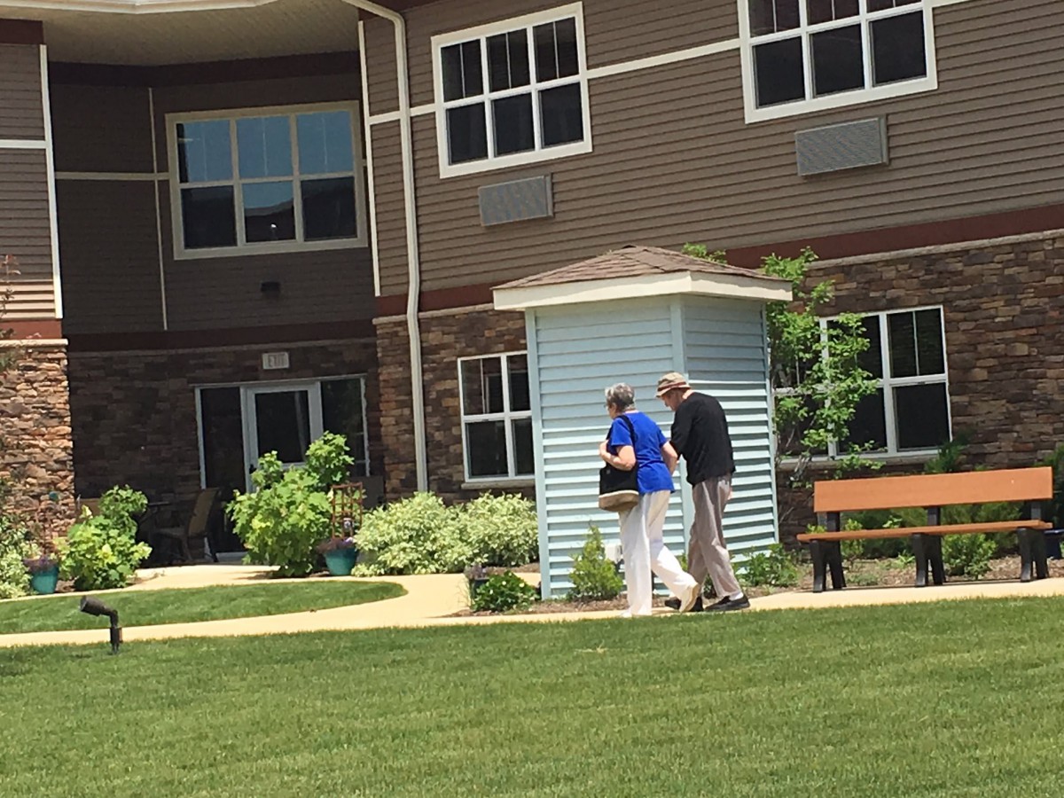 A caregiver assists an elderly person outside a residential building with a garden.