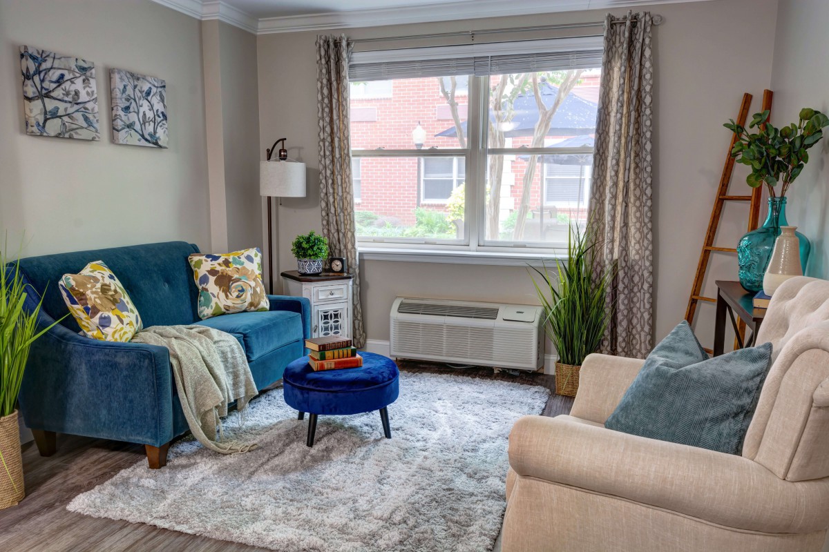 A cozy living room with a blue armchair and beige sofa, featuring modern decor and a large window with curtains.