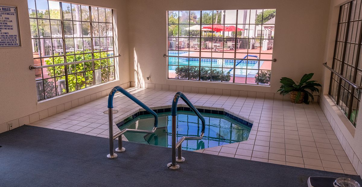 An indoor hot tub with a view of an outdoor swimming pool through large windows.