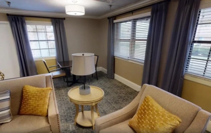 A cozy living room with beige chairs, a round table, lamp, and large windows with curtains.