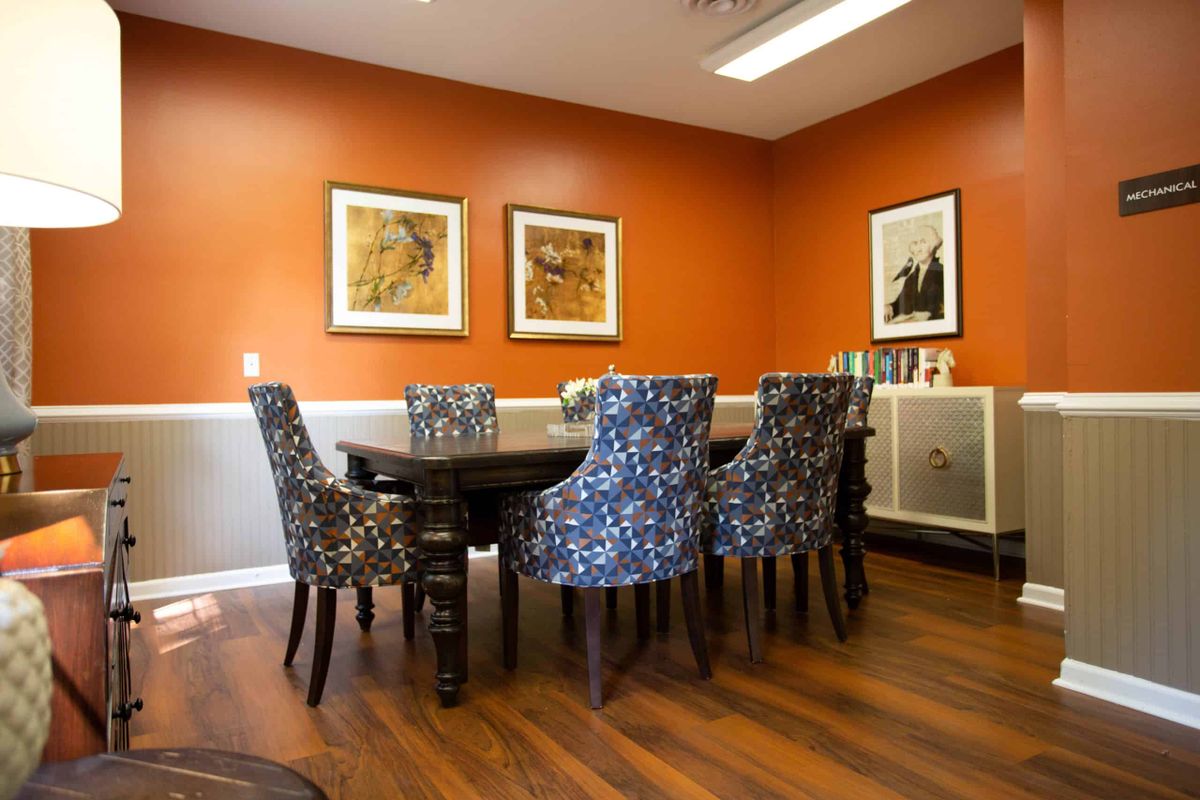 A dining room with an orange accent wall, decorated with framed artwork, featuring a wooden table and patterned chairs on a wooden floor.