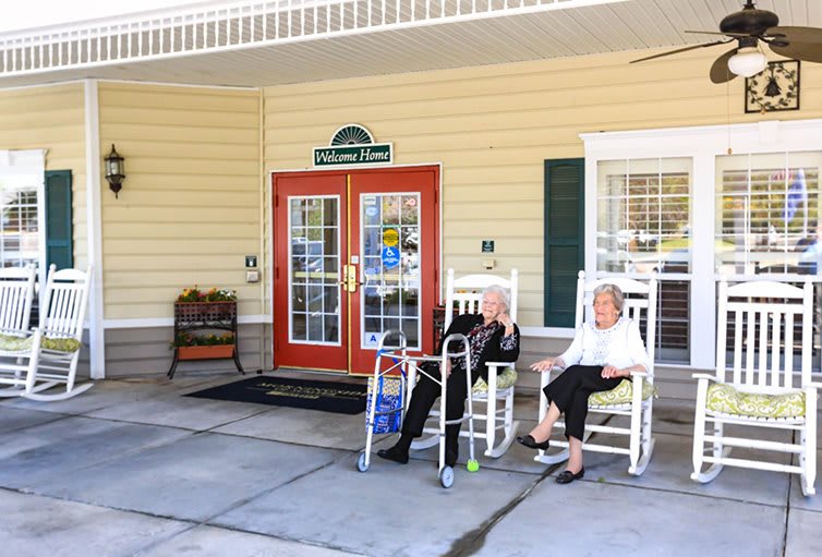 Two elderly individuals relax in rocking chairs on the front porch of a retirement home.