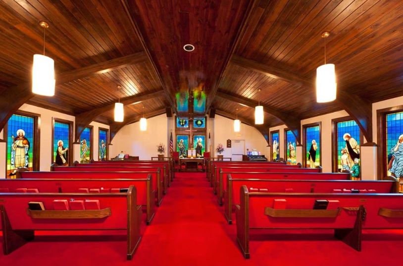 A beautifully lit church interior featuring red carpeted aisles, wooden ceiling, and colorful stained glass windows.