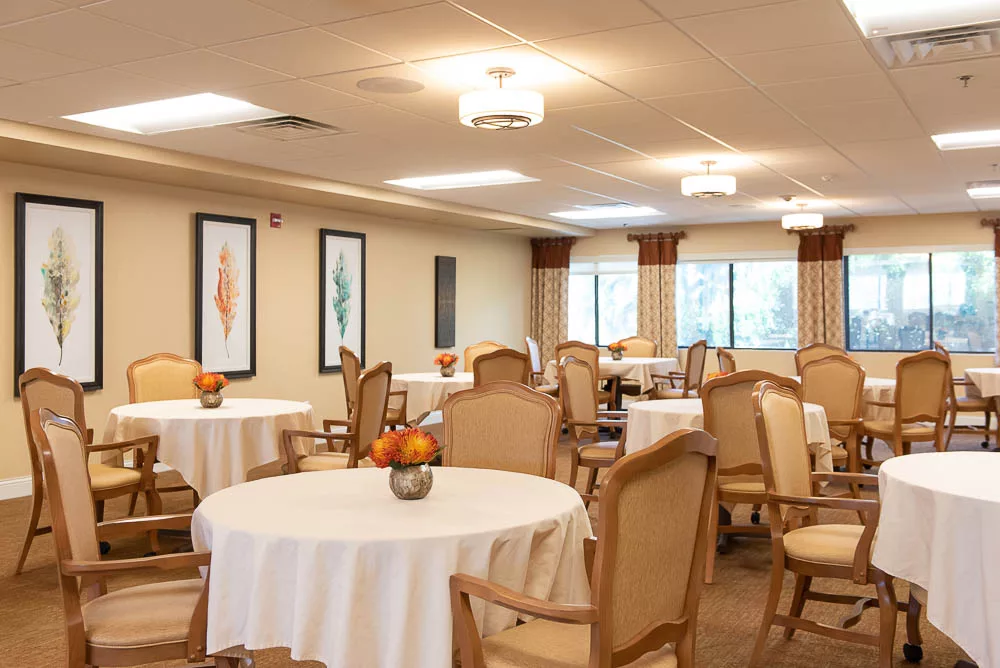A well-lit dining room with round tables and chairs, decorated with framed art on the walls and natural light coming through the windows.
