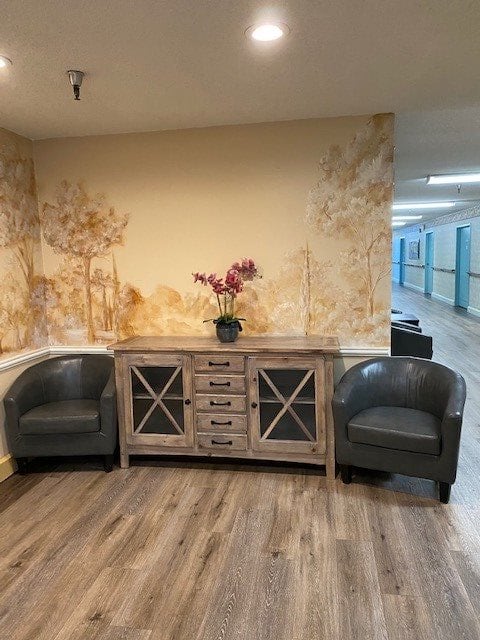 A cozy hallway corner featuring two leather chairs, a wooden cabinet with decorative glass doors, and a potted orchid on top. The backdrop is a mural-style wall art adding warmth to the ambiance.