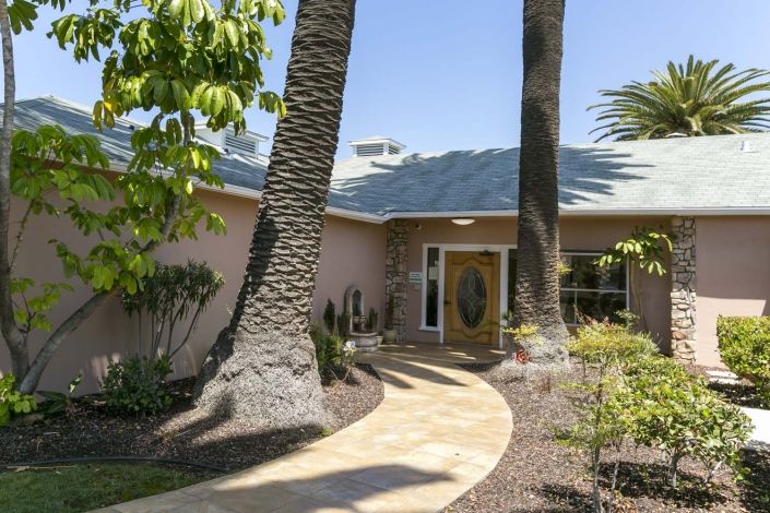 A sunny view of a house entrance with palm trees and a landscaped pathway.