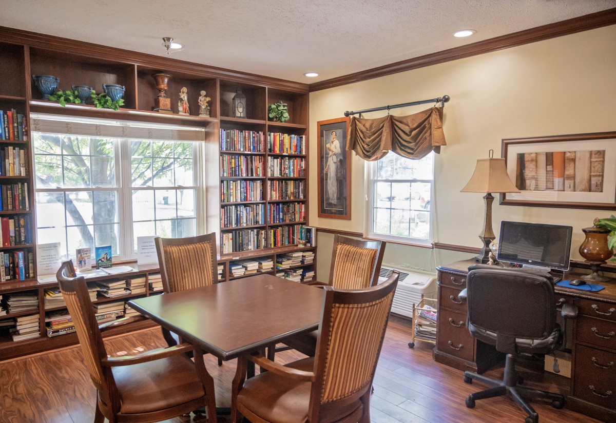 A cozy library room featuring wooden bookshelves, a table with chairs, and a computer by the window.
