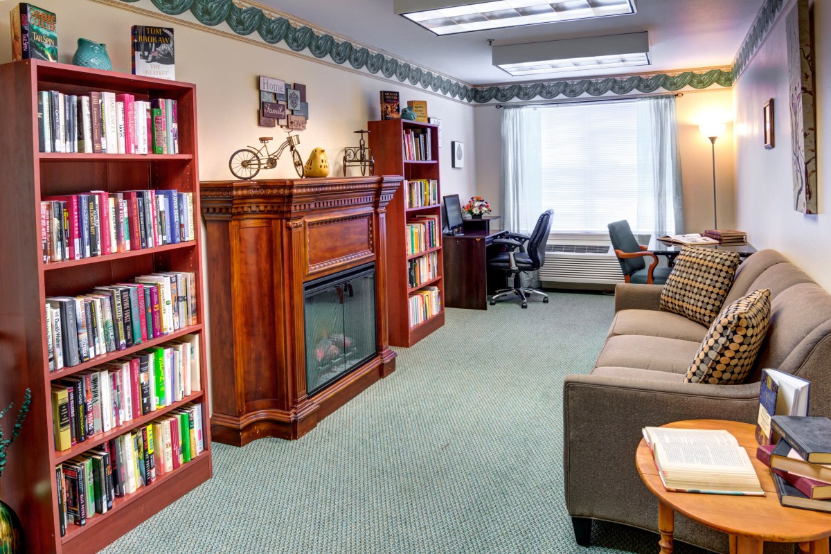 A cozy office space featuring bookshelves, a fireplace, desk, and couch with natural light streaming in through a window.