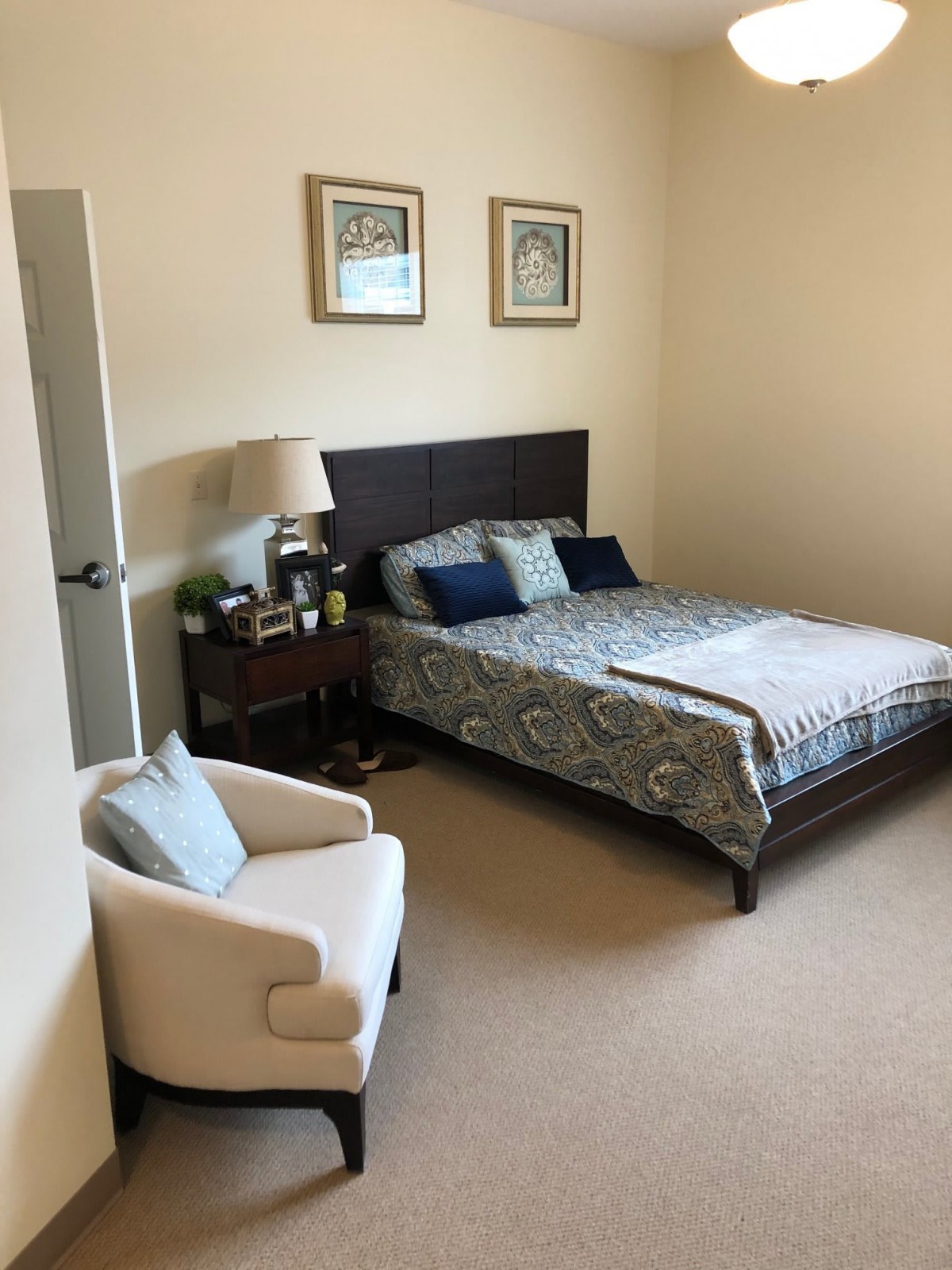 A cozy bedroom with a neatly made bed, featuring decorative pillows and a patterned duvet. A lamp sits on the side table next to the bed, and an armchair is placed in the corner of the room.