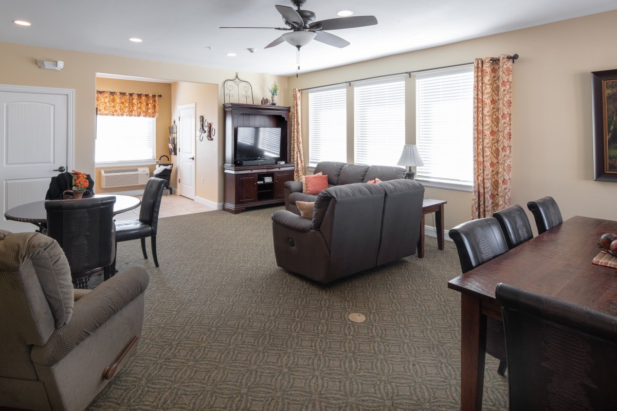 A cozy living room with a sofa set, dining table, and entertainment unit, featuring large windows and a ceiling fan.