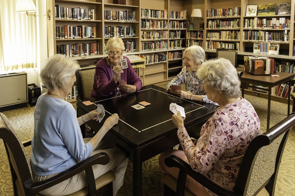A group of seniors playing cards in a library setting surrounded by bookshelves.
