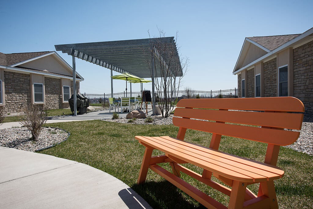 A sunny outdoor area featuring an orange bench, a pergola, and well-maintained green spaces.