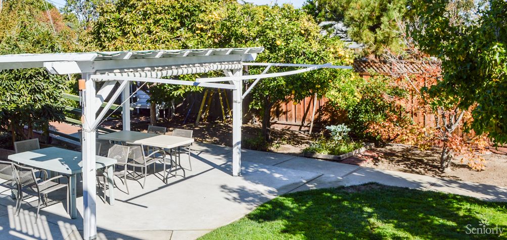 A sunny backyard featuring a pergola over a patio with outdoor furniture, surrounded by lush garden greenery.