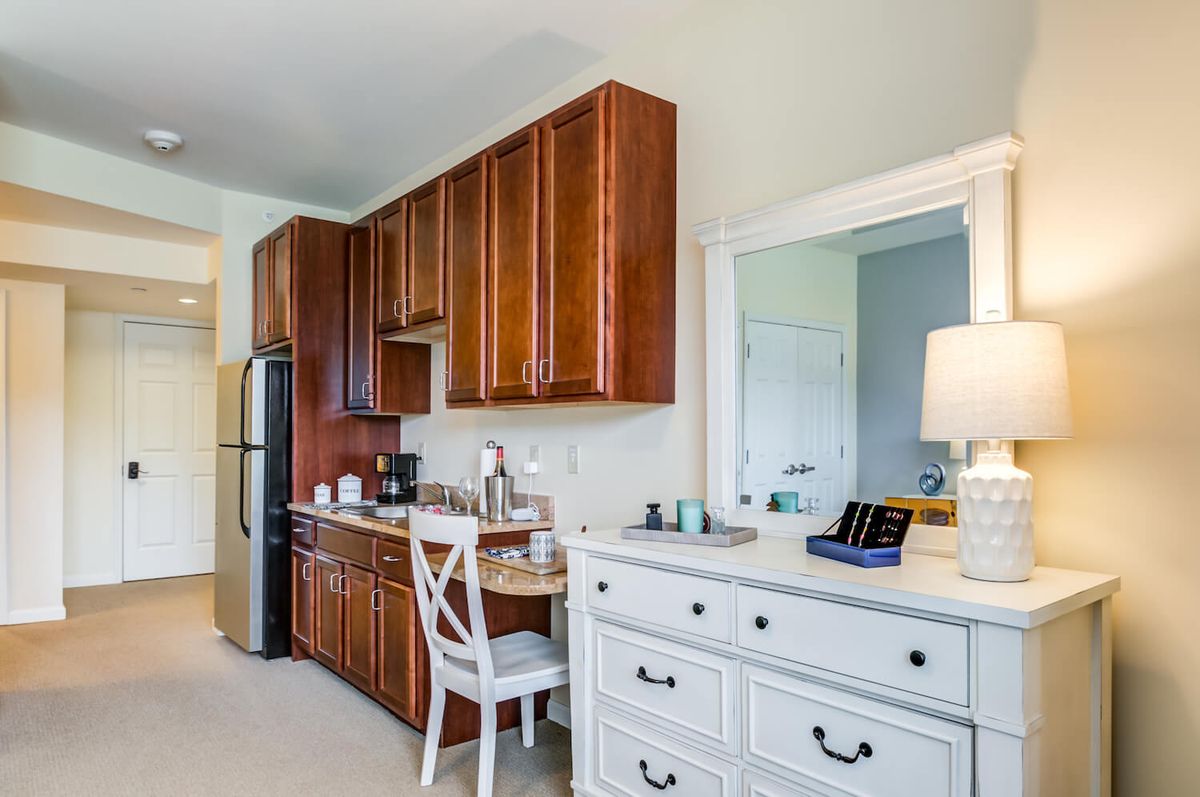 A compact kitchen with wooden cabinets and a white countertop, featuring a small dining area with a chair and a lamp.
