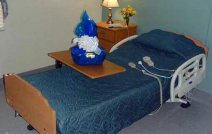 A hospital room featuring a bed with medical equipment, a gift basket on the table, and a lamp on the nightstand.