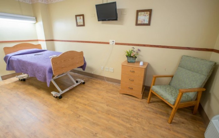 A clean and organized hospital room featuring a bed, a television on the wall, a chair, and a small bedside cabinet.