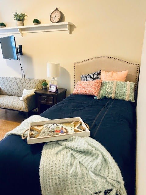 A cozy bedroom with a neatly made bed adorned with colorful pillows and a blanket. A tray is placed on the bed, and there's a lamp on the nightstand next to a small couch.