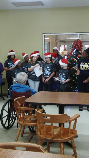 Children in Christmas hats singing for residents at a care facility, spreading holiday cheer.