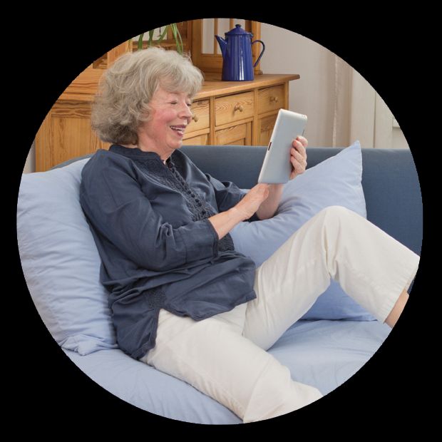 An elderly woman smiling while using a tablet on the couch.