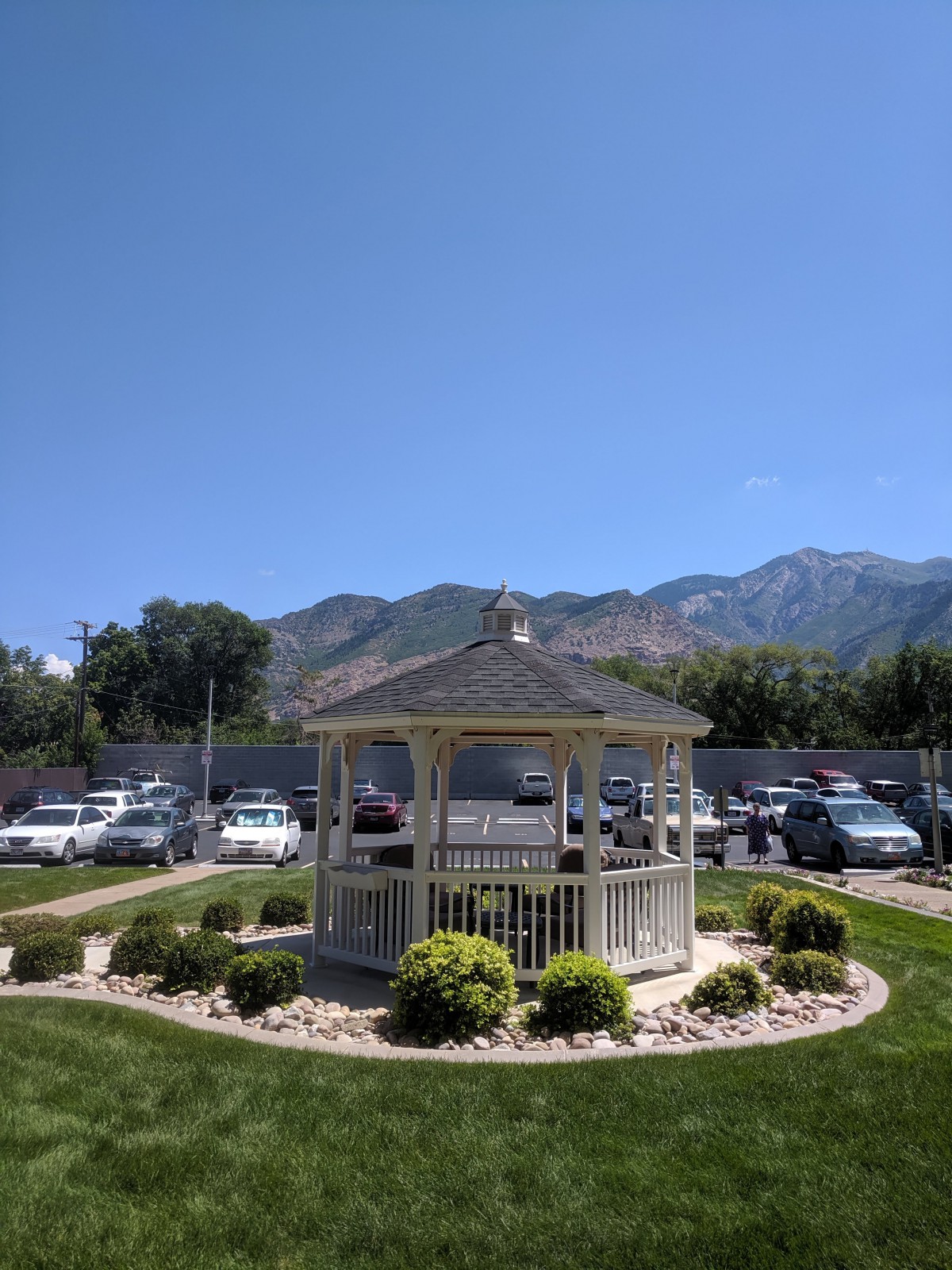 A scenic view of a park with a gazebo, surrounded by parked cars and mountains under a clear blue sky.