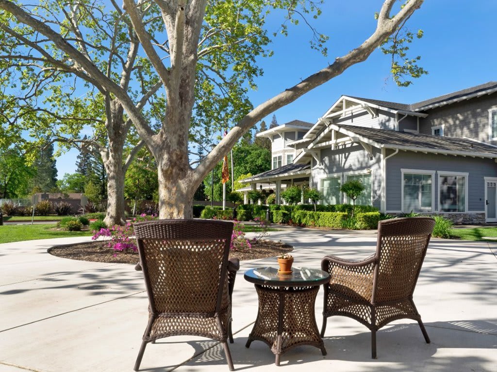 A tranquil patio setting with wicker chairs and a table overlooking a garden and house on a sunny day.