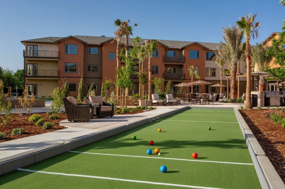 A well-maintained bocce court adjacent to a residential building with patio seating and lush landscaping.