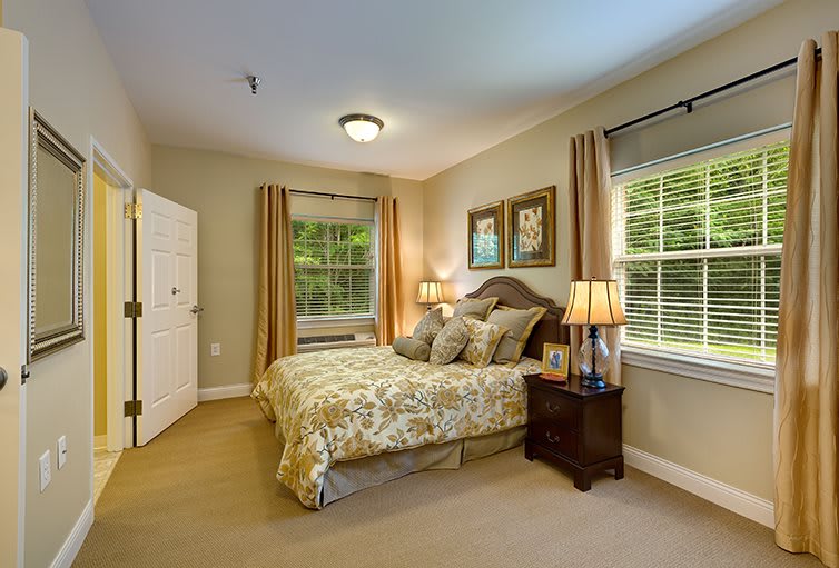 A cozy bedroom featuring a large bed with decorative pillows, bedside tables with lamps, and wall art. The room has carpeted flooring and is well-lit by natural light through the windows.