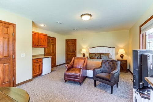 A spacious bedroom with armchairs, a bed, wooden cabinets, and warm lighting.