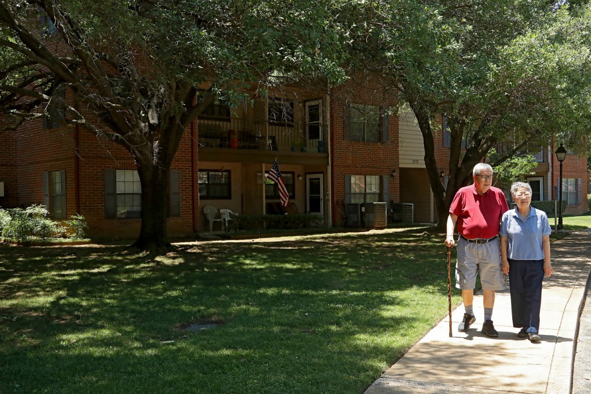 A man using a cane walks on a sidewalk in front of an apartment building on a sunny day.