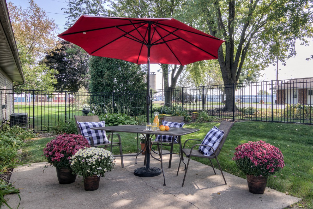A cozy outdoor patio setup with a red umbrella, table, and chairs surrounded by potted flowers.