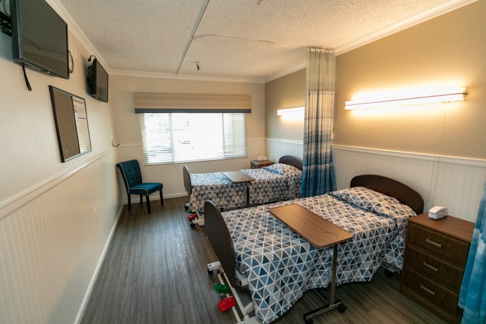 A hospital room with two beds, divided by a curtain, featuring overhead lighting and a window.